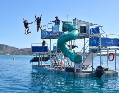 People jumping off a water park platform with a green spiral slide and multiple levels, set in a tropical ocean environment under a bright blue sky.