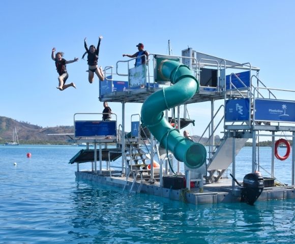 People jumping off a water park platform with a green spiral slide and multiple levels, set in a tropical ocean environment under a bright blue sky.