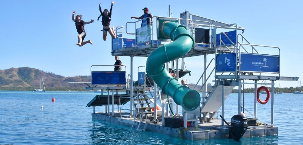 People jumping off a water park platform with a green spiral slide and multiple levels, set in a tropical ocean environment under a bright blue sky.