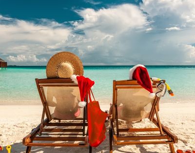 Two beach chairs facing a turquoise ocean, with a starry hat and Santa hats draped over them, evoke a festive, tropical holiday vibe.