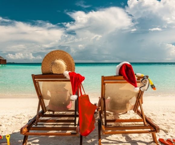 Two beach chairs facing a turquoise ocean, with a starry hat and Santa hats draped over them, evoke a festive, tropical holiday vibe.