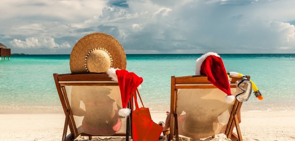 Two beach chairs facing a turquoise ocean, with a starry hat and Santa hats draped over them, evoke a festive, tropical holiday vibe.