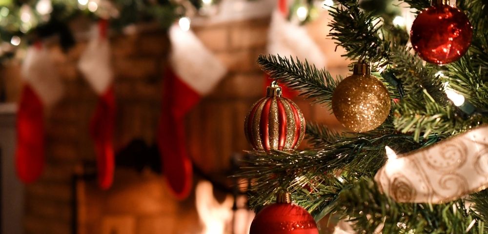 Close-up of a decorated Christmas tree with red and gold ornaments beside a lit fireplace. Stockings hang on the mantel, evoking a cozy, festive atmosphere.