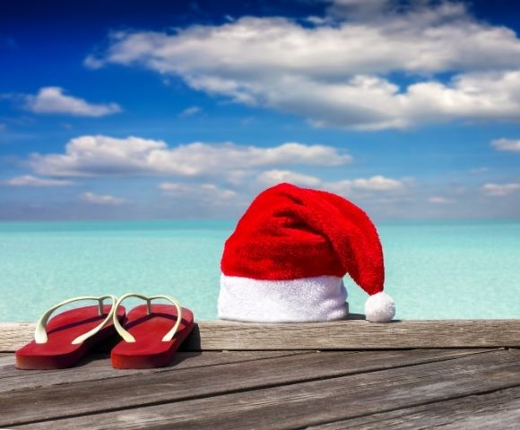 Red flip-flops and a Santa hat rest on a wooden dock overlooking a turquoise ocean under a blue sky with scattered clouds, evoking tropical holiday vibes.