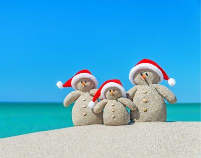 Three sand snowmen with red Santa hats stand on a beach. They have pebble eyes, carrot noses, and button details, against a vivid blue sky and turquoise sea.