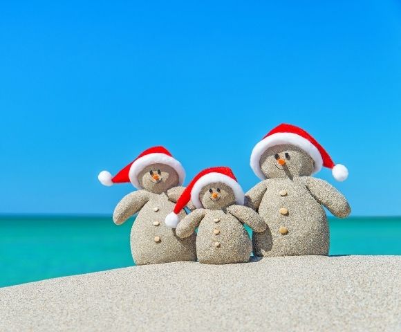 Three sand snowmen with red Santa hats stand on a beach. They have pebble eyes, carrot noses, and button details, against a vivid blue sky and turquoise sea.