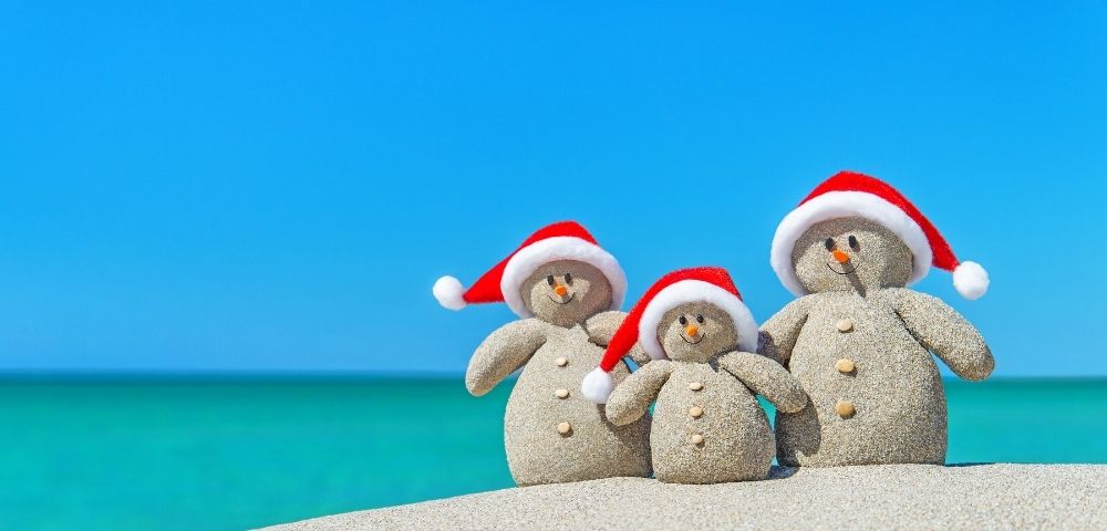 Three sand snowmen with red Santa hats stand on a beach. They have pebble eyes, carrot noses, and button details, against a vivid blue sky and turquoise sea.