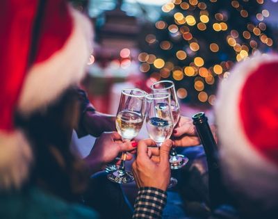 A festive gathering with people in Santa hats clinking champagne glasses. A blurred Christmas tree with twinkling lights sets a joyful tone.