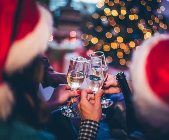 A festive gathering with people in Santa hats clinking champagne glasses. A blurred Christmas tree with twinkling lights sets a joyful tone.