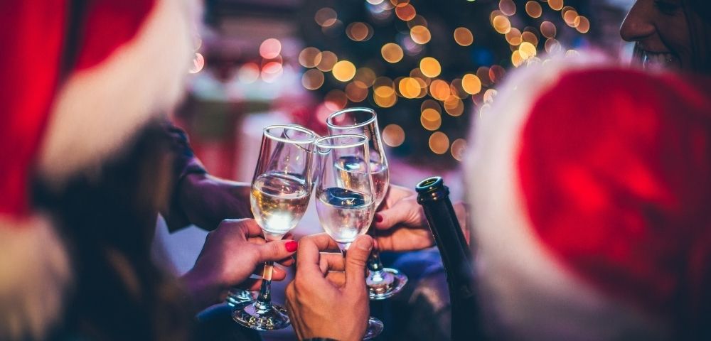 A festive gathering with people in Santa hats clinking champagne glasses. A blurred Christmas tree with twinkling lights sets a joyful tone.