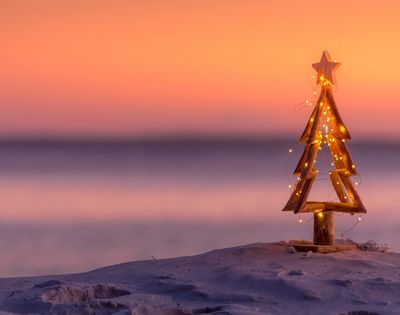 Wooden Christmas tree adorned with string lights, set on a sandy beach. A serene sunset paints the sky in warm hues of orange and pink, evoking tranquility.