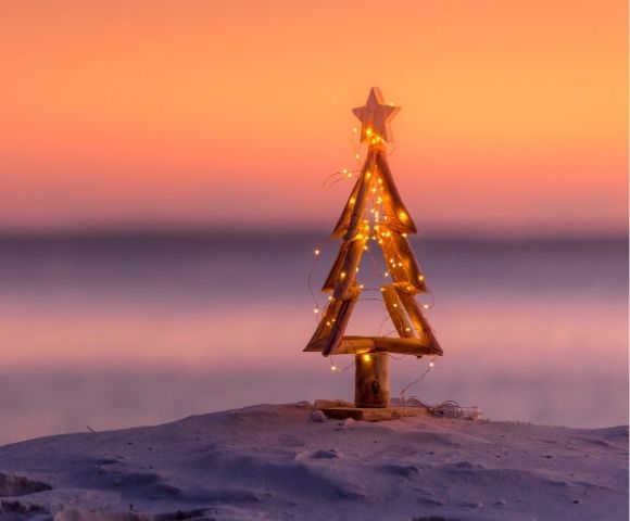 Wooden Christmas tree adorned with string lights, set on a sandy beach. A serene sunset paints the sky in warm hues of orange and pink, evoking tranquility.