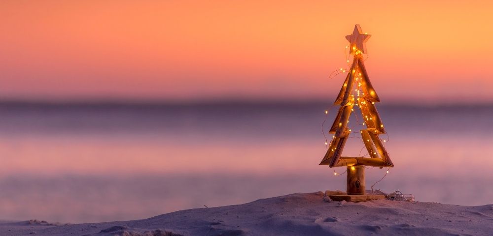 Wooden Christmas tree adorned with string lights, set on a sandy beach. A serene sunset paints the sky in warm hues of orange and pink, evoking tranquility.