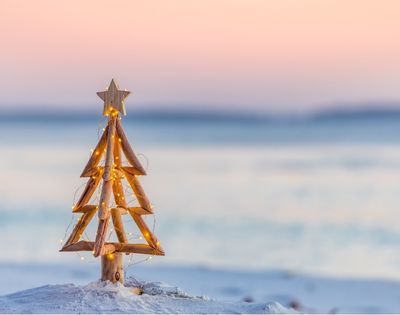 Wooden Christmas tree with a star topper stands on sandy beach against a bright blue ocean backdrop. The scene is serene and summery.