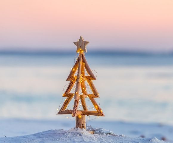 Wooden Christmas tree with a star topper stands on sandy beach against a bright blue ocean backdrop. The scene is serene and summery.
