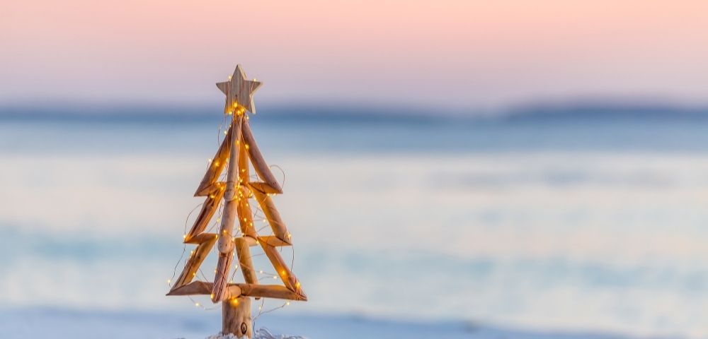 Wooden Christmas tree with a star topper stands on sandy beach against a bright blue ocean backdrop. The scene is serene and summery.