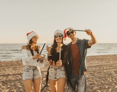 Three people wearing Santa hats and sunglasses celebrate on a beach at sunset, holding sparklers and a drink, creating a joyful holiday atmosphere.