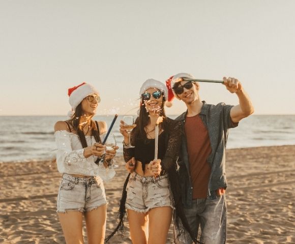 Three people wearing Santa hats and sunglasses celebrate on a beach at sunset, holding sparklers and a drink, creating a joyful holiday atmosphere.
