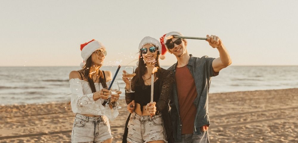 Three people wearing Santa hats and sunglasses celebrate on a beach at sunset, holding sparklers and a drink, creating a joyful holiday atmosphere.