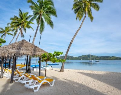 Idyllic beach scene featuring palm trees, a thatched hut, yellow lounge chairs, and calm waters with boats in the distance.