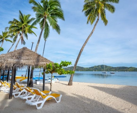 Idyllic beach scene featuring palm trees, a thatched hut, yellow lounge chairs, and calm waters with boats in the distance.
