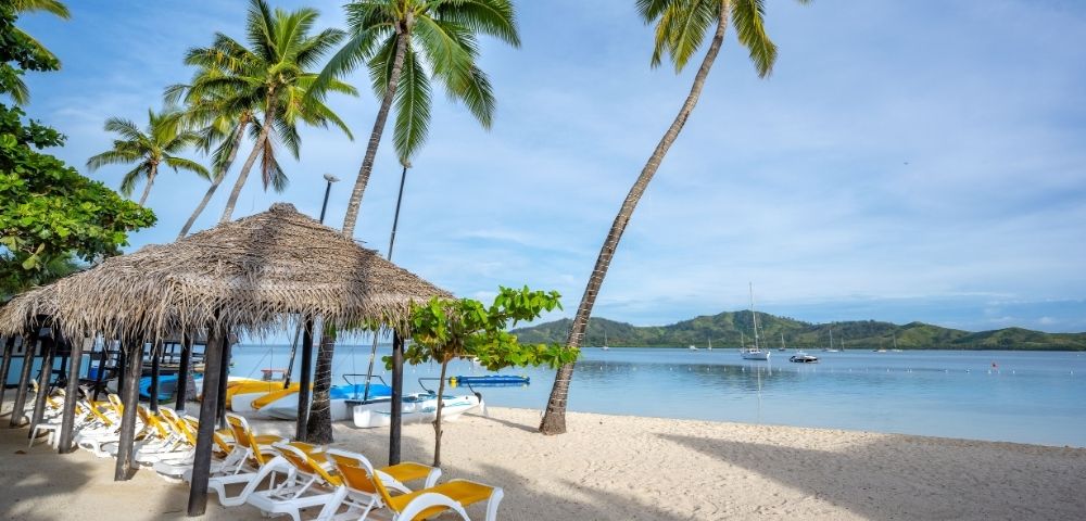 Idyllic beach scene featuring palm trees, a thatched hut, yellow lounge chairs, and calm waters with boats in the distance.