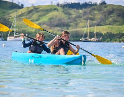 Two individuals paddle a blue kayak with yellow paddles on a calm, clear water, surrounded by green hills and boats in the background.