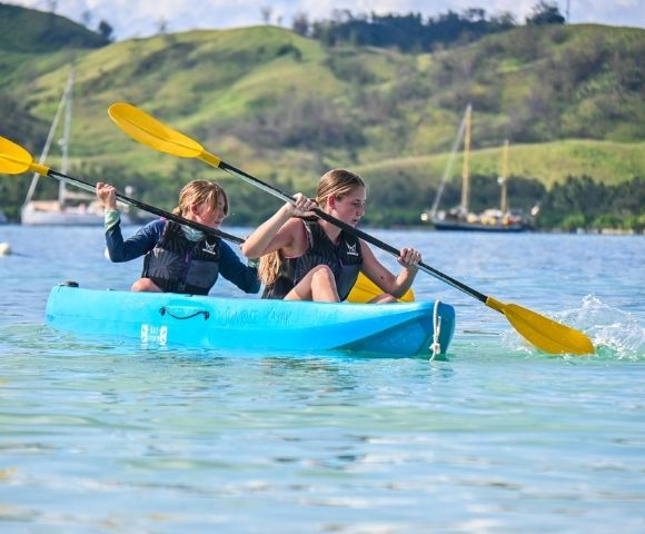 Two individuals paddle a blue kayak with yellow paddles on a calm, clear water, surrounded by green hills and boats in the background.