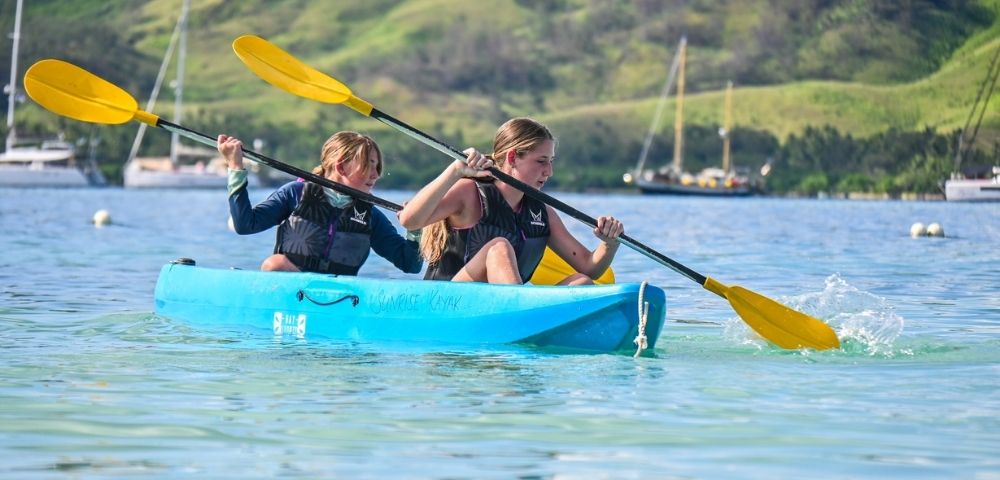 Two individuals paddle a blue kayak with yellow paddles on a calm, clear water, surrounded by green hills and boats in the background.