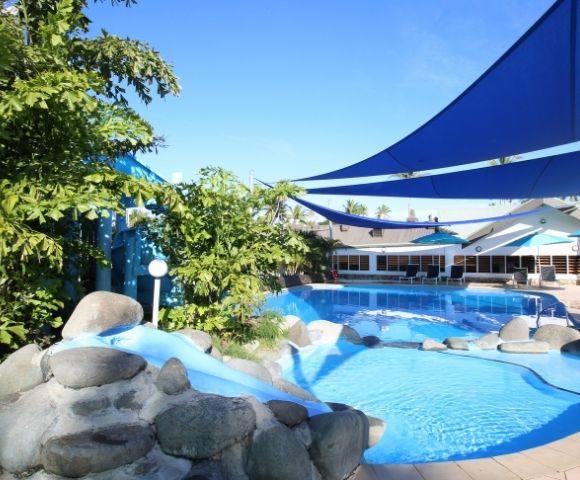 An outdoor swimming pool with a small slide and large boulders, under blue sunshades, with a resort building in the background under a clear blue sky.