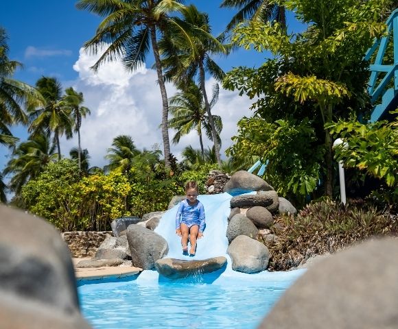 A young child in a blue swimsuit slides down a small, blue water slide into a turquoise pool, surrounded by rocks, greenery, and palm trees under a bright blue sky with fluffy white clouds.