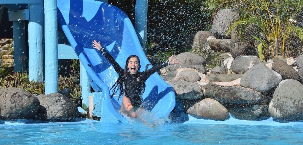 A young girl with long dark hair wearing a black rash guard and green bottoms slides down a blue water slide with her arms outstretched, water splashing, and a joyful expression.