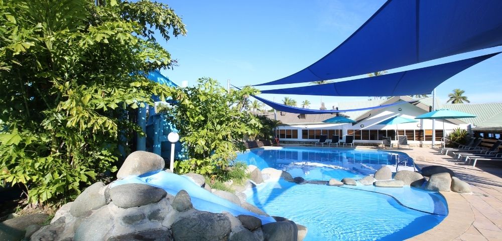 An outdoor swimming pool with a small slide and large boulders, under blue sunshades, with a resort building in the background under a clear blue sky.