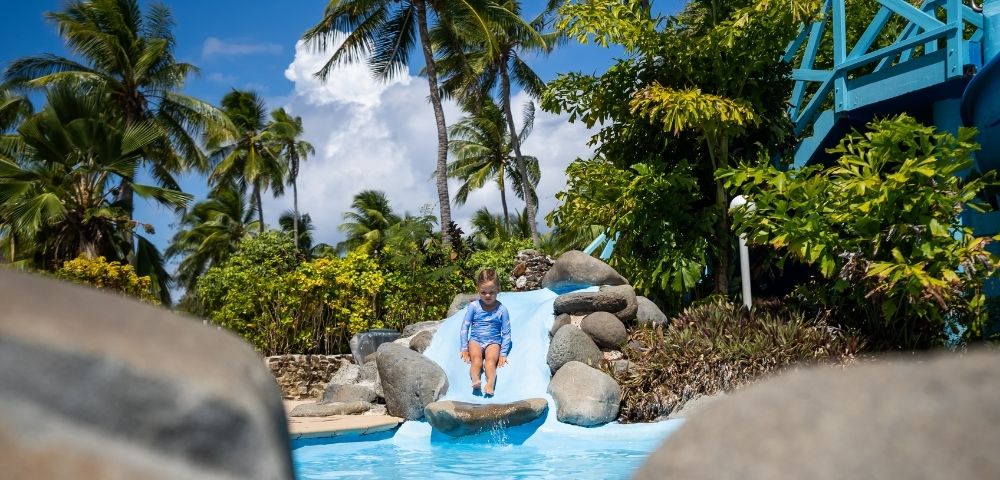A young child in a blue swimsuit slides down a small, blue water slide into a turquoise pool, surrounded by rocks, greenery, and palm trees under a bright blue sky with fluffy white clouds.