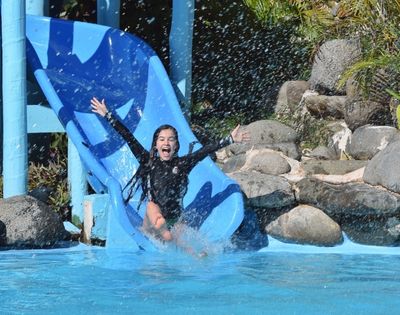 A young girl with long dark hair wearing a black rash guard and green bottoms slides down a blue water slide with her arms outstretched, water splashing, and a joyful expression.