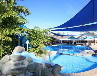 An outdoor swimming pool with a small slide and large boulders, under blue sunshades, with a resort building in the background under a clear blue sky.