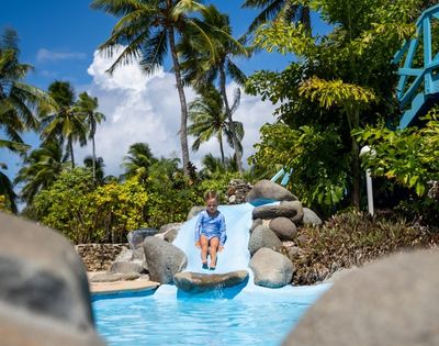 A young child in a blue swimsuit slides down a small, blue water slide into a turquoise pool, surrounded by rocks, greenery, and palm trees under a bright blue sky with fluffy white clouds.