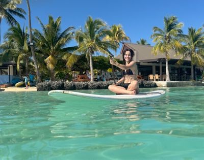 A smiling woman in a black bikini sits on a white paddleboard in clear turquoise water, holding a paddle, with a tropical beach and palm trees in the background.