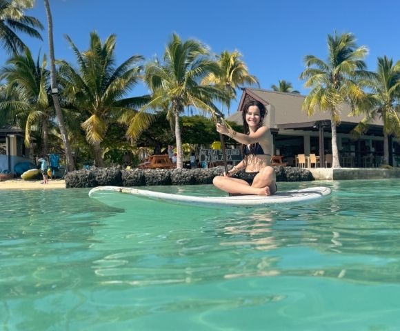 A smiling woman in a black bikini sits on a white paddleboard in clear turquoise water, holding a paddle, with a tropical beach and palm trees in the background.