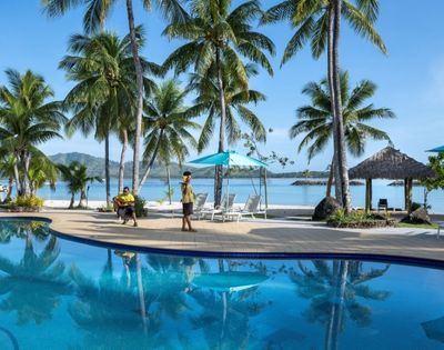 An outdoor shot of a tropical resort featuring a large blue swimming pool reflecting palm trees, with white sand beach in the background, blue ocean, and a clear sky. Two people are near the pool: one playing guitar and another in a uniform carrying a tray. Several palm trees, seating, and beach umbrellas dot the landscape.