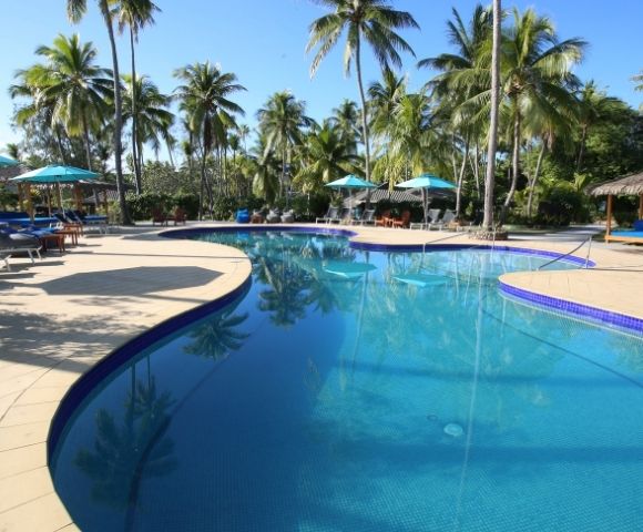 A turquoise-tiled swimming pool curves through a tropical resort setting, surrounded by a beige tiled deck, palm trees, and shaded lounge areas.