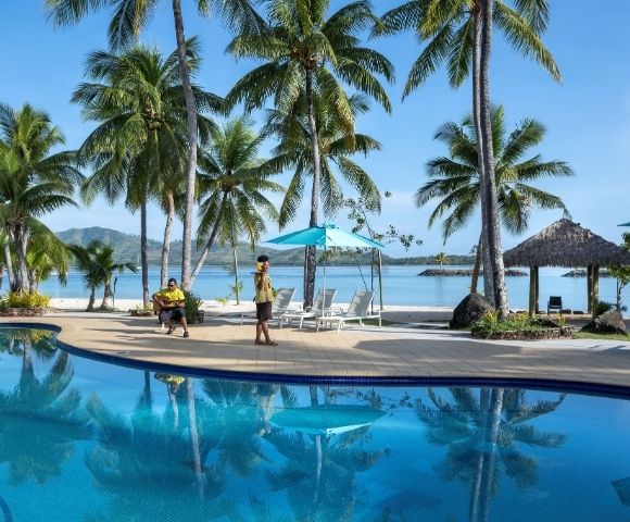 An outdoor shot of a tropical resort featuring a large blue swimming pool reflecting palm trees, with white sand beach in the background, blue ocean, and a clear sky. Two people are near the pool: one playing guitar and another in a uniform carrying a tray. Several palm trees, seating, and beach umbrellas dot the landscape.