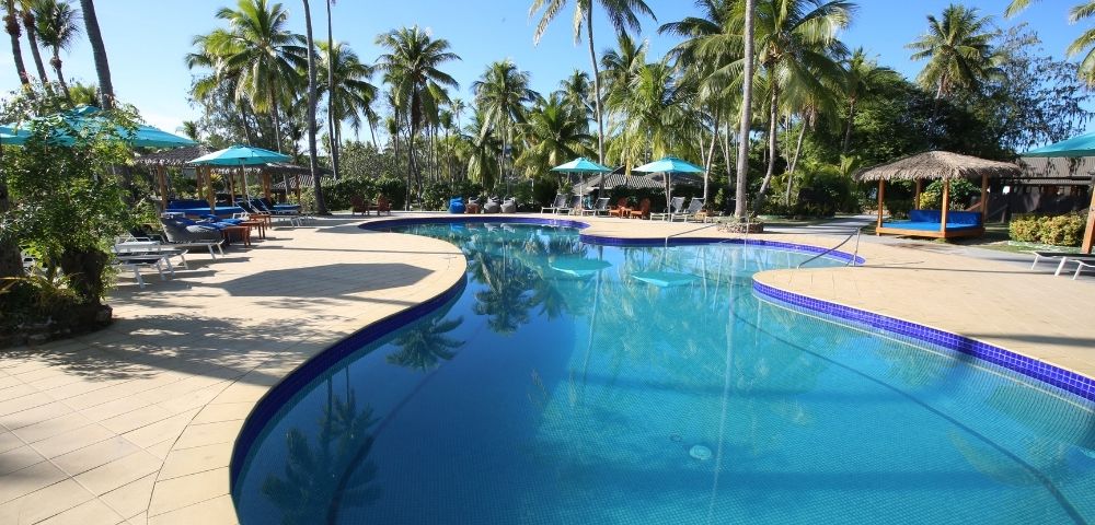 A turquoise-tiled swimming pool curves through a tropical resort setting, surrounded by a beige tiled deck, palm trees, and shaded lounge areas.