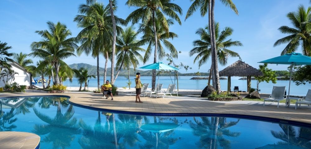 An outdoor shot of a tropical resort featuring a large blue swimming pool reflecting palm trees, with white sand beach in the background, blue ocean, and a clear sky. Two people are near the pool: one playing guitar and another in a uniform carrying a tray. Several palm trees, seating, and beach umbrellas dot the landscape.