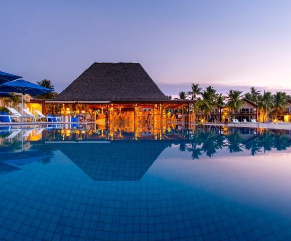 A luxury resort at dusk, featuring a pool reflecting the lit bar and palm trees, with lounge chairs and blue umbrellas.