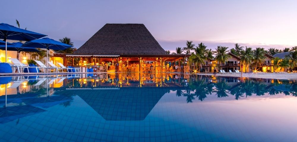 A luxury resort at dusk, featuring a pool reflecting the lit bar and palm trees, with lounge chairs and blue umbrellas.