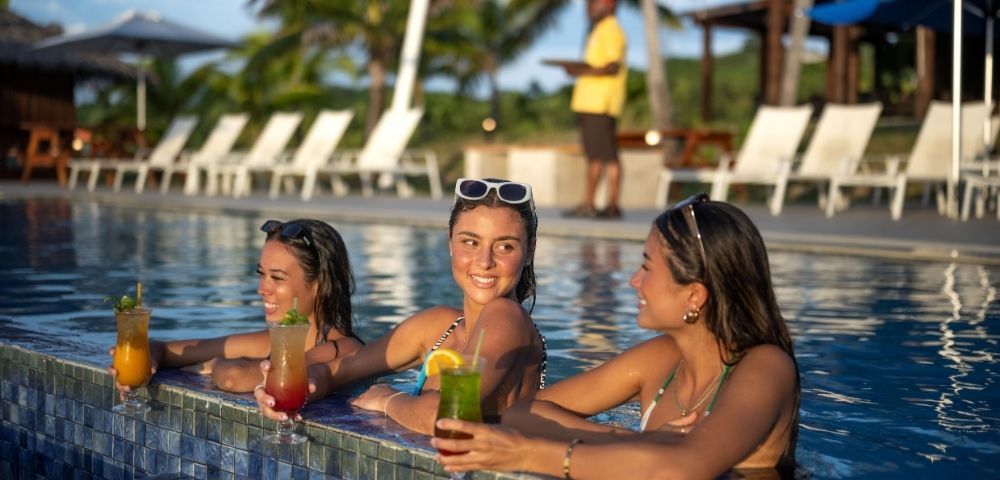 Three young women in swimsuits, each holding a cocktail, lean on the edge of a blue-tiled swimming pool at a resort, with a server carrying a tray in the background.