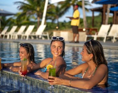 Three young women in swimsuits, each holding a cocktail, lean on the edge of a blue-tiled swimming pool at a resort, with a server carrying a tray in the background.