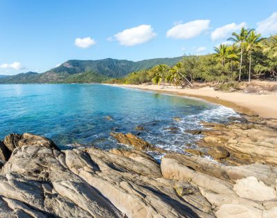 Rocky coastline with turquoise ocean waves, sandy beach, and lush palm trees under a clear blue sky. The scene conveys tranquility and natural beauty.