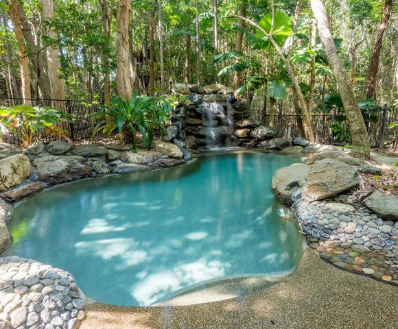 Tranquil natural pool with a waterfall, surrounded by rocks and lush greenery in a forest setting. Sunlight filters through the trees, creating a serene atmosphere.